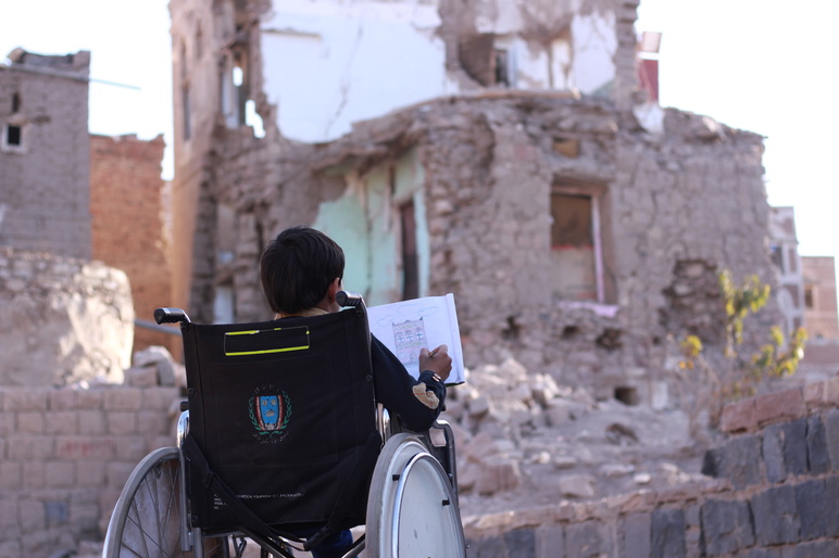 A Yemeni boy in a wheelchair draws in front of a destroyed house